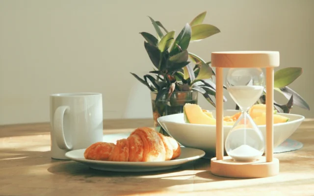 Mesa com alimentos naturais, café e ampulheta iluminadas por luz natural, simbolizando o tempo e o conhecimento como formas de nutrição.