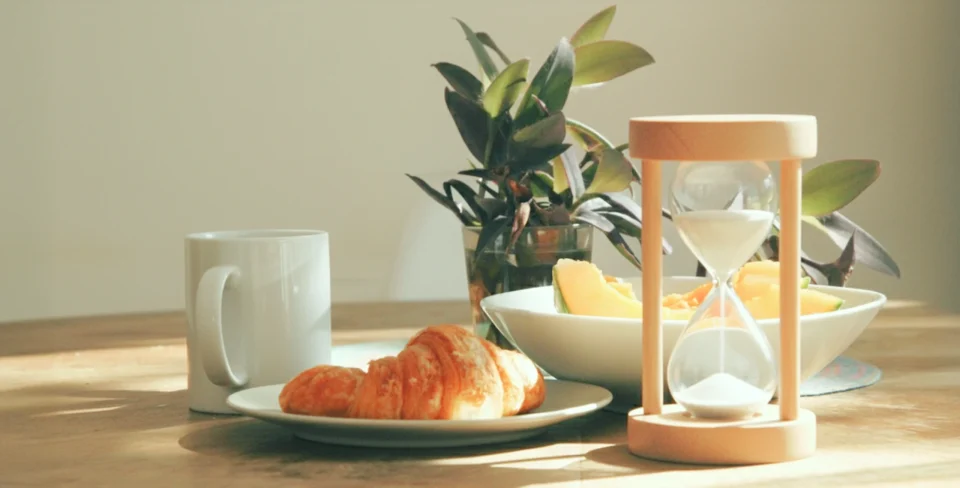 Mesa com alimentos naturais, café e ampulheta iluminadas por luz natural, simbolizando o tempo e o conhecimento como formas de nutrição.